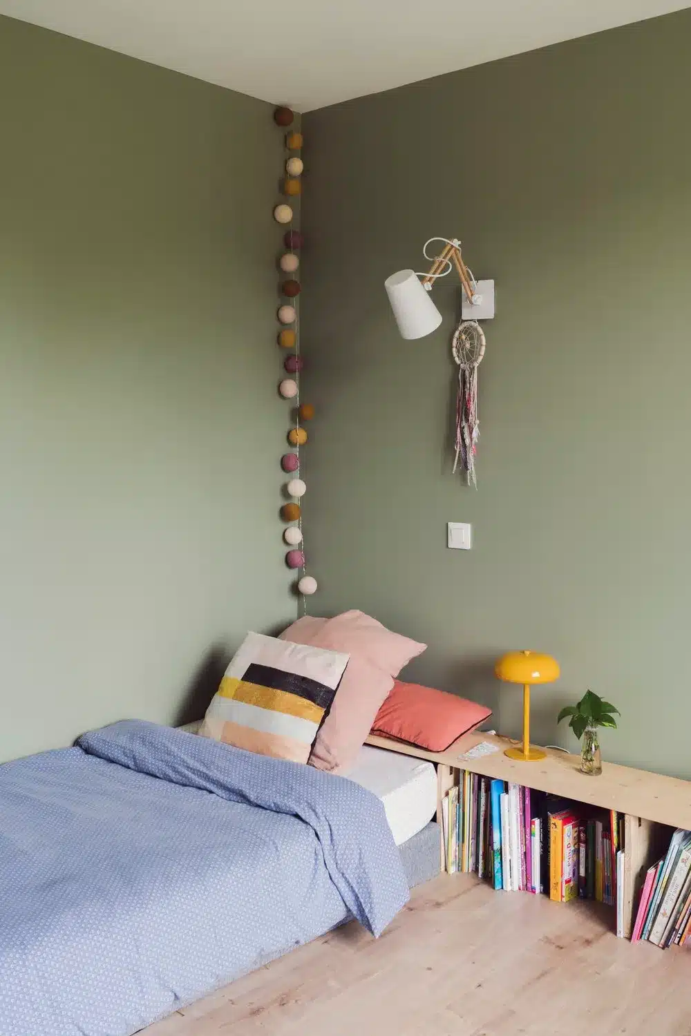 Chambre d'enfant dans un joli ton vert lichen, un long meuble de rangement qui accueille les livres et de jolis draps douillets.