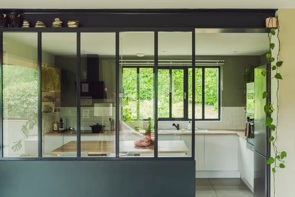 Photo d'une verrière atelier séparant un séjour d'une cuisine blanche et bois avec murs verts.