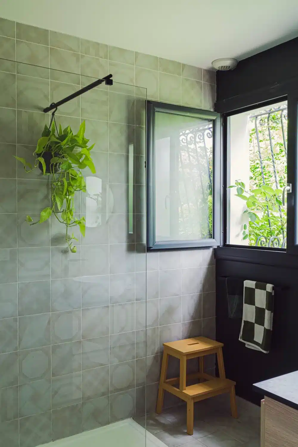 Photo d'une salle de bain au mur noir associé à des carreaux de ciment, le tout relevé d'un tabouret jaune et d'une plante qui cascade dans la douche vitrée.