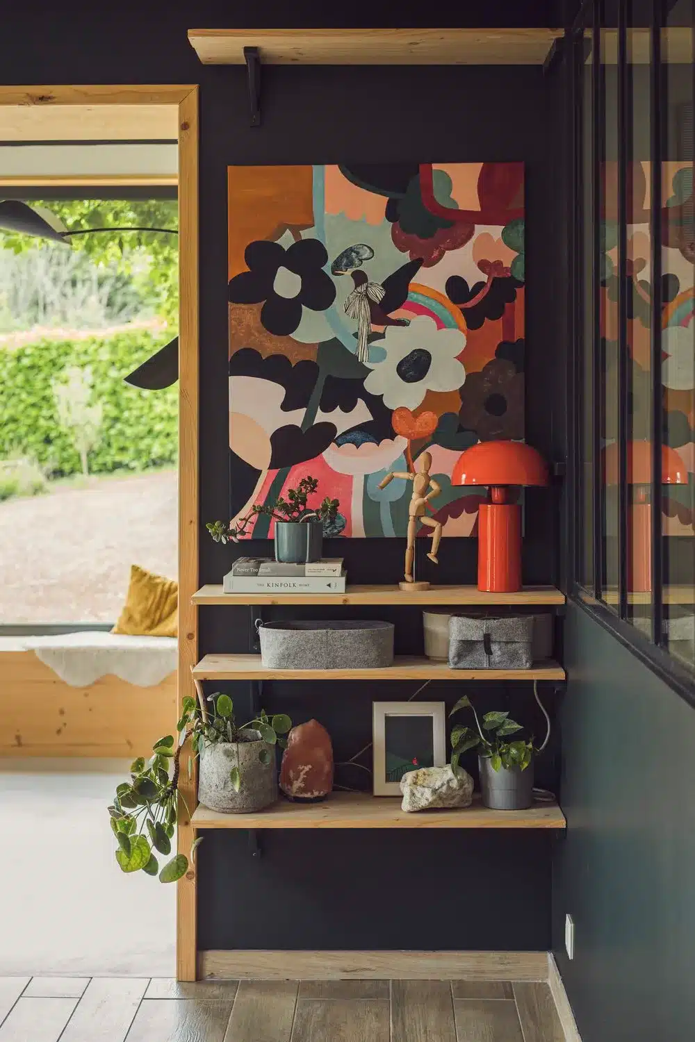 Photo d'étagères en ois contre un mur vert foncé avec beaucoup de caractère. les touches de couleurs du tableau peint à la main et de la petite lampe orange vibrent et l'oeil est attiré vers l'extérieur en second plan.